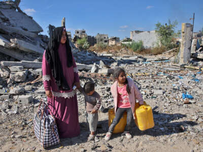 A woman and her two children carry water and a single bag of their belongings amidst destroyed buildings