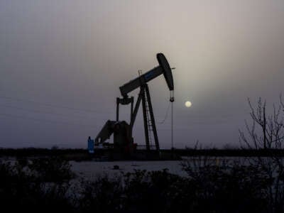 A pumpjack is seen against a grey sky and a hazy sun