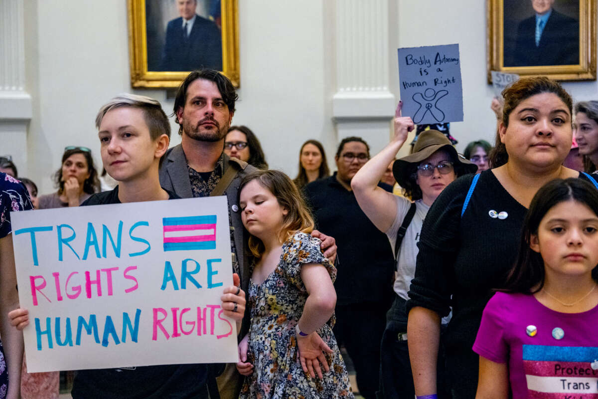 People gathered inside of a governmental building display signs, one of which reads "TRANS RIGHTS ARE HUMAN RIGHTS"