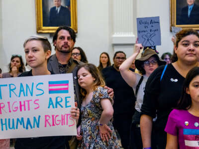 People gathered inside of a governmental building display signs, one of which reads "TRANS RIGHTS ARE HUMAN RIGHTS"