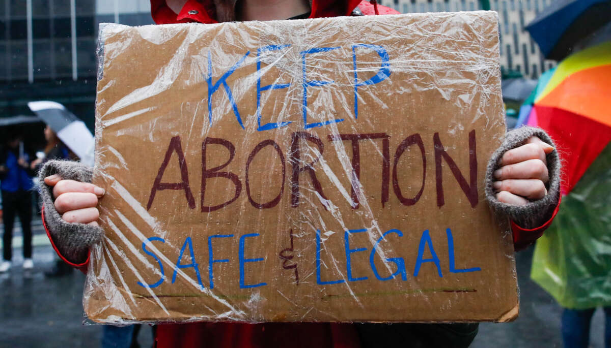 A person holds a cardboard sign coverfed in plastic reading "KEEP ABORTION SAFE & LEGAL" during an outdoor protest in the rain