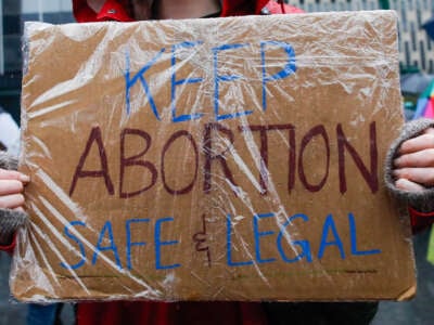 A person holds a cardboard sign coverfed in plastic reading "KEEP ABORTION SAFE & LEGAL" during an outdoor protest in the rain