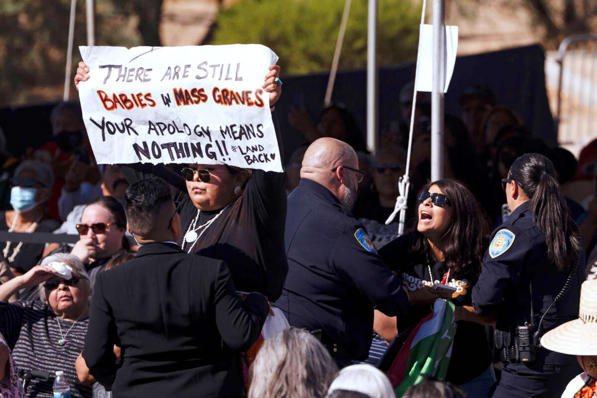 A woman holding a sign reading "THERE ARE STILL BABIES IN MASS GRAVES; YOUR APOLOGY MEANS NOTHING #landback" is harassed by police