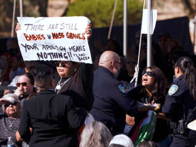 A woman holding a sign reading "THERE ARE STILL BABIES IN MASS GRAVES; YOUR APOLOGY MEANS NOTHING #landback" is harassed by police