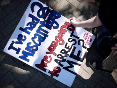 A person writes a protest sign reading "I'VE HAD 2 MISCARRIAGES; ARE YOU GOING TO ARREST ME TOO? #FUABBOT #DOBETTERTEXAS" on the ground