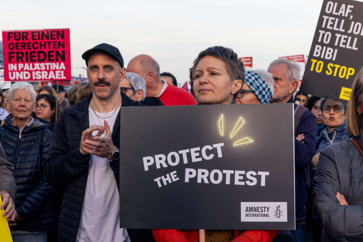 A protester holding a sign reading "PROTECT THE PROTEST" stands beside others, one with a sign in German, and another with a sign reading "OLAF, TELL JOE TO TELL BIBI TO STOP"