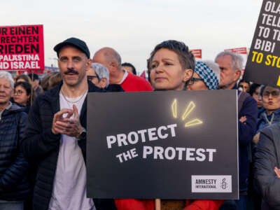 A protester holding a sign reading "PROTECT THE PROTEST" stands beside others, one with a sign in German, and another with a sign reading "OLAF, TELL JOE TO TELL BIBI TO STOP"