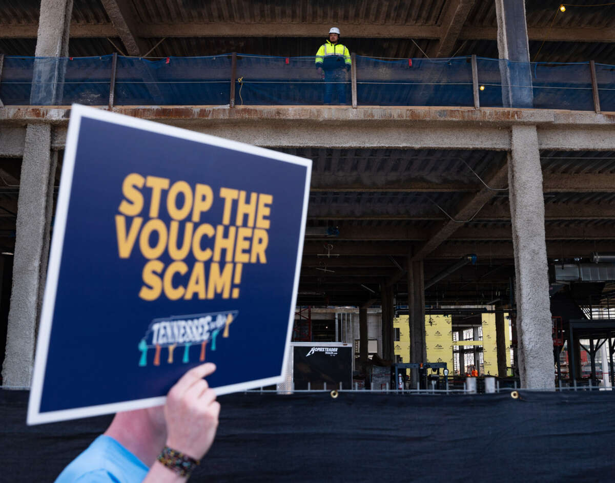 A protester holds up a sign reading "STOP THE VOUCHER SCAM" while construction workers on a nearby project site look on