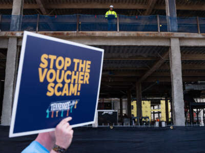 A protester holds up a sign reading "STOP THE VOUCHER SCAM" while construction workers on a nearby project site look on