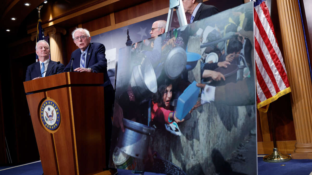 Bernie Sanders stands at a podium alongside other senators and an enlarged photo of hungry Palestinian children struggling to get food