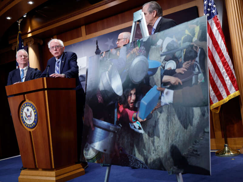 Bernie Sanders stands at a podium alongside other senators and an enlarged photo of hungry Palestinian children struggling to get food