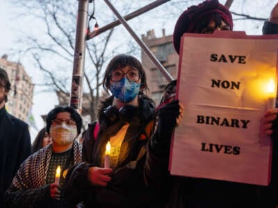 People wearing masks and holding electric candles participate in a vigil as one holds a sign reading "SAVE NON BINARY LIVES"