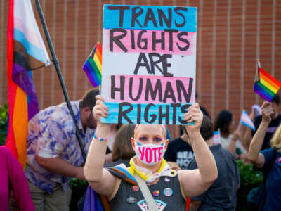 A protester in a mask that reads "VOTE" holds up a sign reading "TRANS RIGHTS ARE HUMAN RIGHTS" during an outdoor demonstration