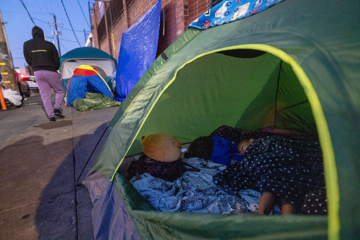 Children in a green tent on the sidewalk as an adult walks past them in the distance