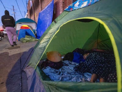 Children in a green tent on the sidewalk as an adult walks past them in the distance