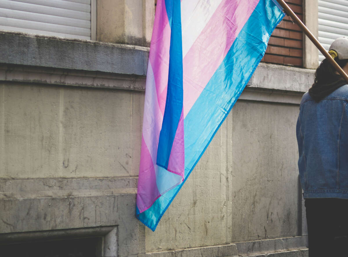 Person holds a transgender flag by the side of a building.