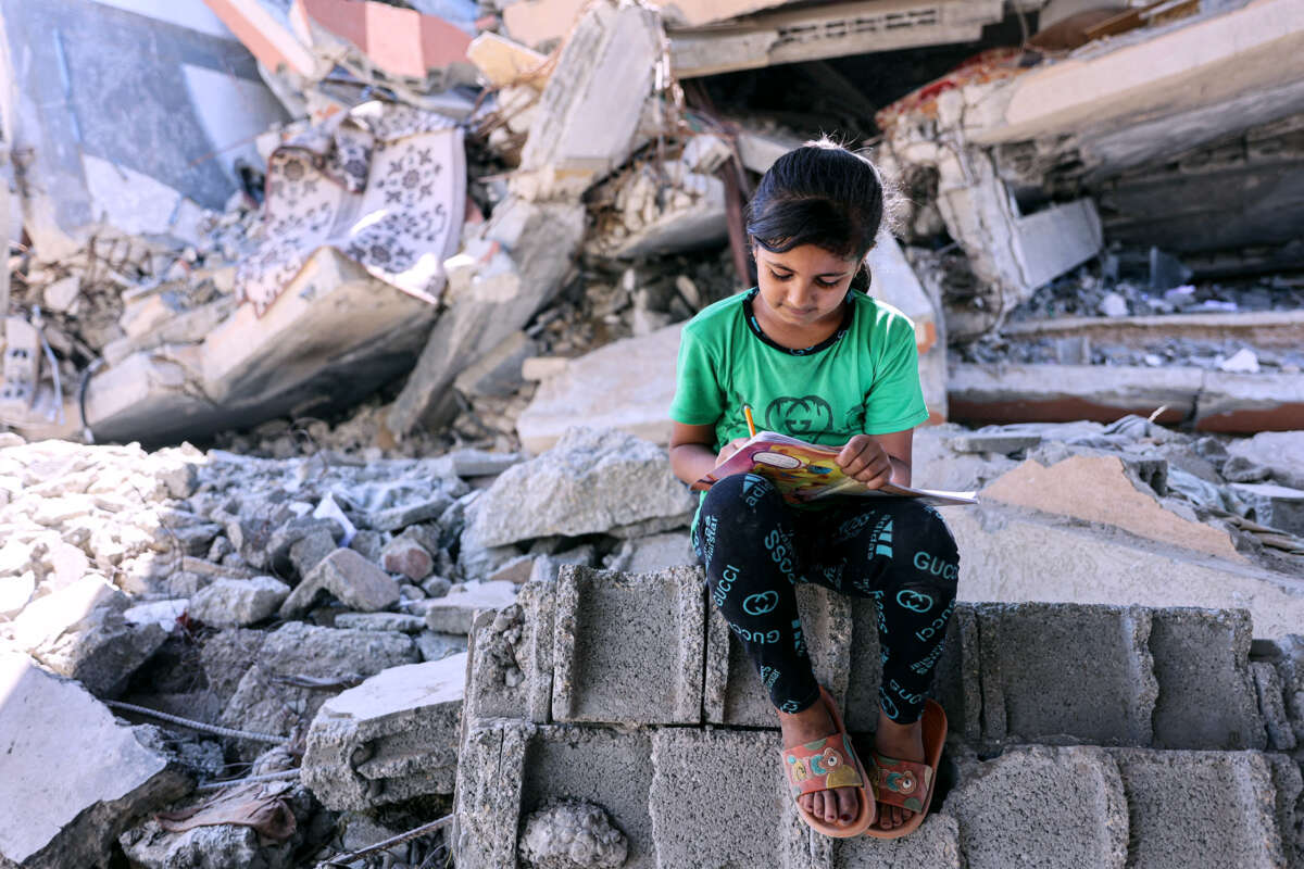 A girl in a green shirt writes in a notebook while seated atop rubble from homes and businesses destroyed by the IOF