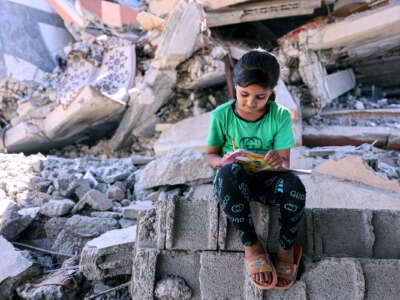 A girl in a green shirt writes in a notebook while seated atop rubble from homes and businesses destroyed by the IOF