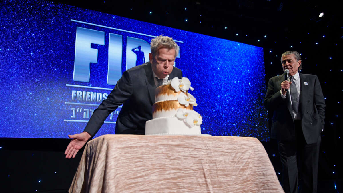 A man blows on a cake bearing no visible candles while standing in front of a sign reading "FIDF"