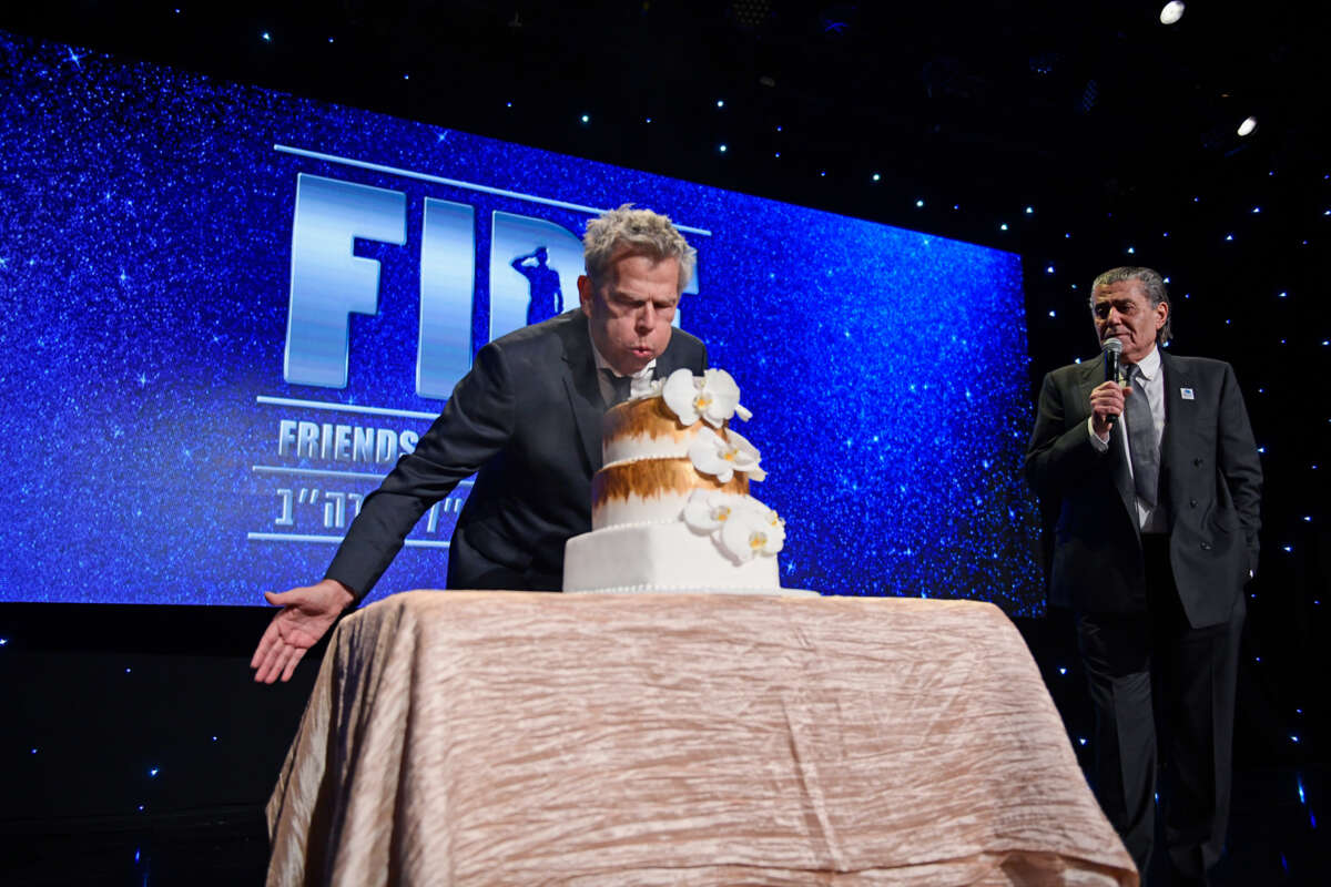 A man blows on a cake bearing no visible candles while standing in front of a sign reading "FIDF"