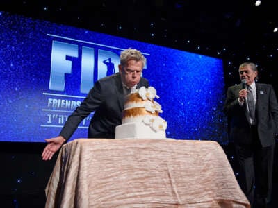 A man blows on a cake bearing no visible candles while standing in front of a sign reading "FIDF"