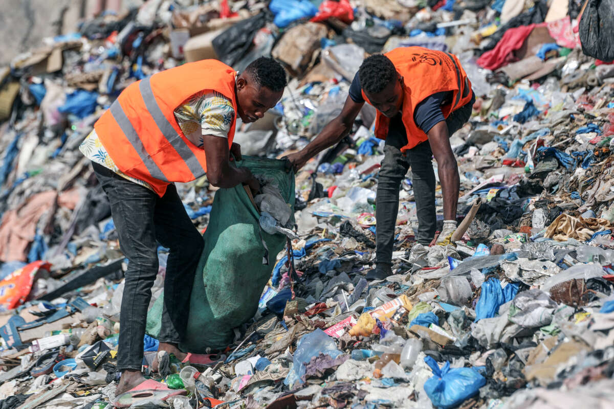 Two men in orange vests clean a beach covered in garbage