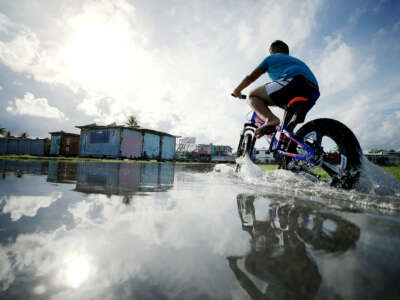 A boy rides his bike through floodwaters towards the sun, the partially-cloudy sky reflected in the water below him