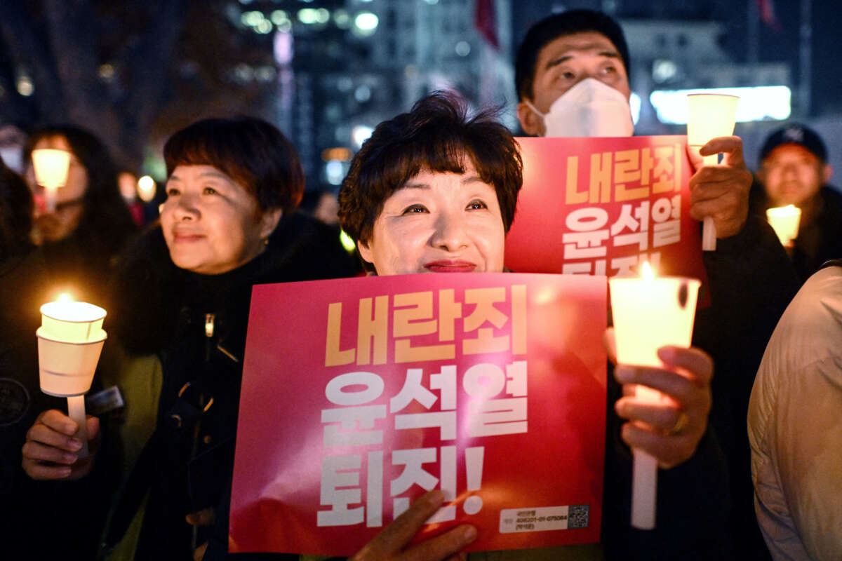 A protester holding a sign reading "Insurrection Yoon Suk Yeol step down!" in Korean smiles while holding a candle