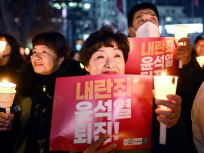 A protester holding a sign reading "Insurrection Yoon Suk Yeol step down!" in Korean smiles while holding a candle