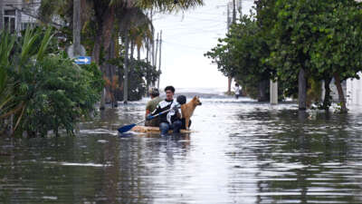 A man paddles himself, two other people and a dog through floodwaters on a raft made out of storm debris