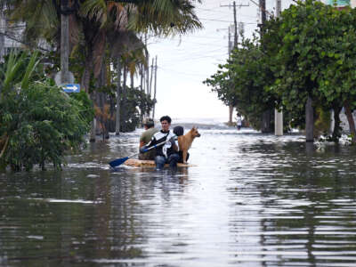 A man paddles himself, two other people and a dog through floodwaters on a raft made out of storm debris