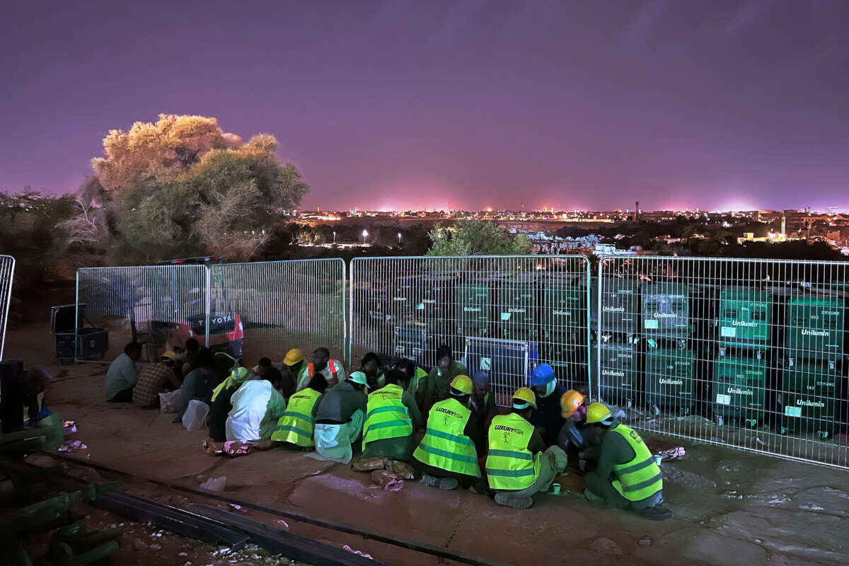 Migrant workers in bright yellow vests huddle together on the ground as a bright skyline lights the sky behind them