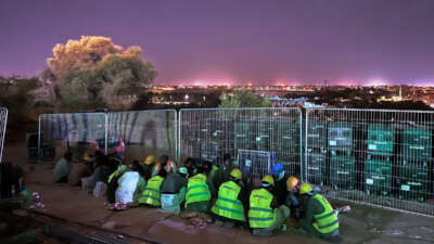 Migrant workers in bright yellow vests huddle together on the ground as a bright skyline lights the sky behind them