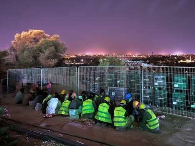 Migrant workers in bright yellow vests huddle together on the ground as a bright skyline lights the sky behind them