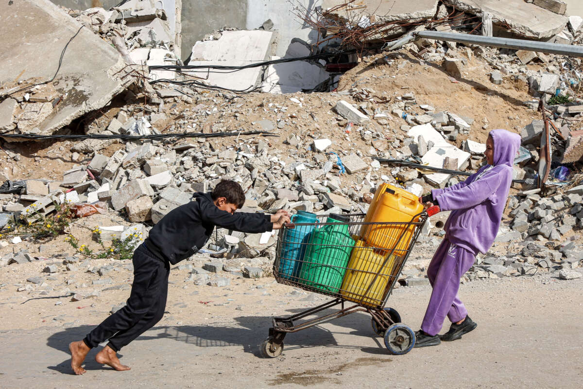 A girl pulls while a boy pushes a shopping cart loaded with filled-up water containers past a mound of rubble and debris in Gaza City on December 11, 2024.