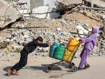 A girl pulls while a boy pushes a shopping cart loaded with filled-up water containers past a mound of rubble and debris in Gaza City on December 11, 2024.