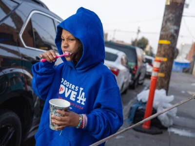 A little girl in a blue hoodie brushes her teeth with a disposable cup while standing on the sidewalk