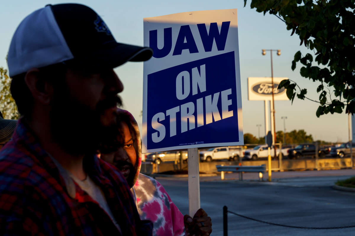 Two people stand near eachother while one holds a sign reading "UAW ON STRIKE" during an outdoor strike