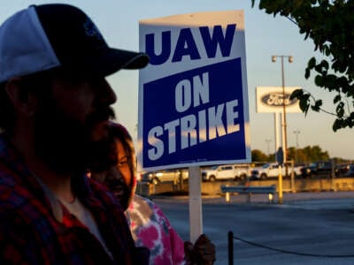 Two people stand near eachother while one holds a sign reading "UAW ON STRIKE" during an outdoor strike