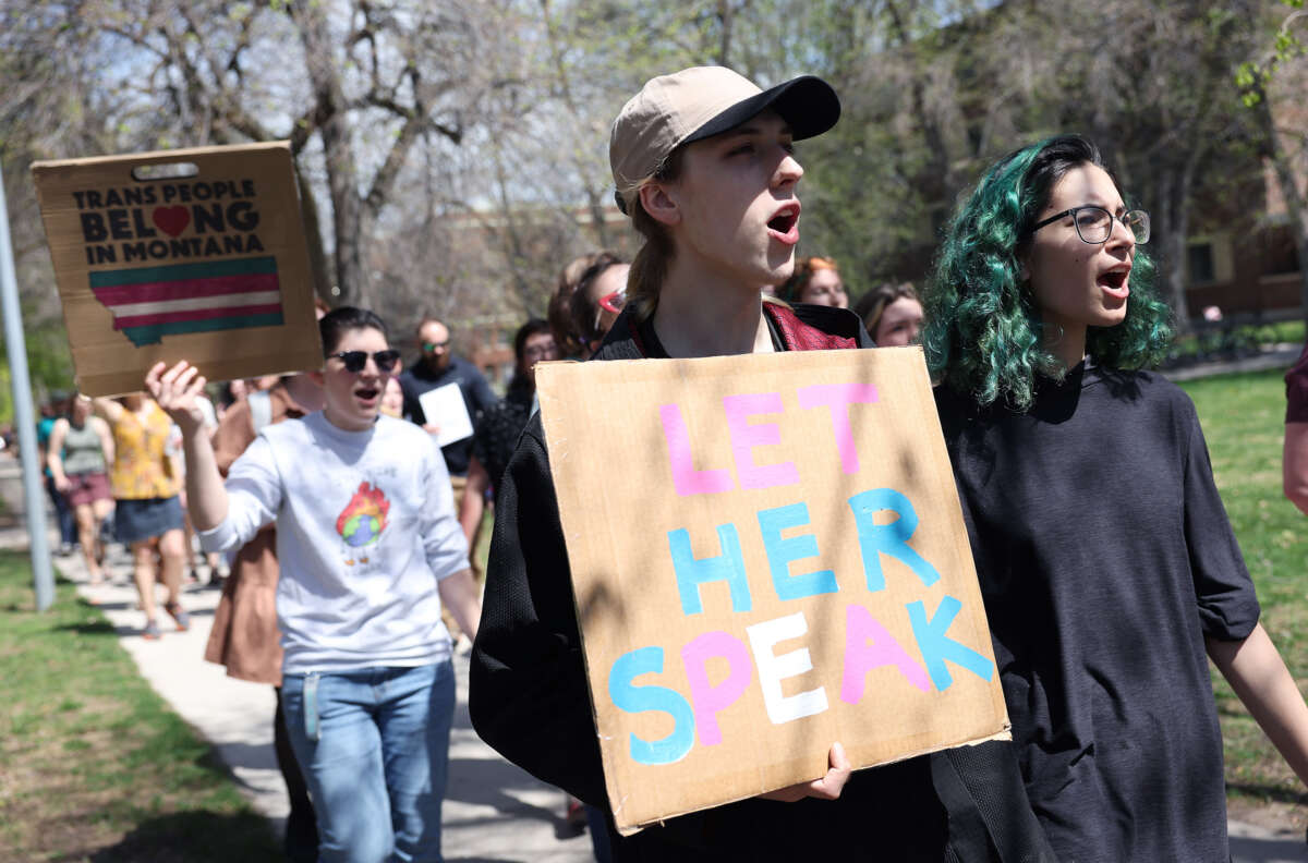 A person holding a cardboard sign reading "LET HER SPEAK" in pink, blue and white letters is joined by others in an outdoor protest