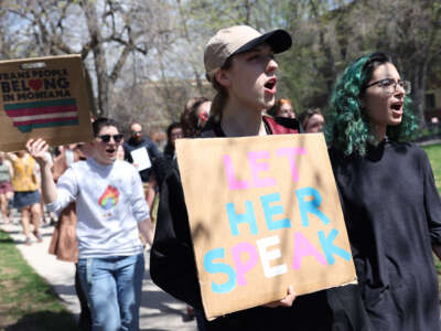 A person holding a cardboard sign reading "LET HER SPEAK" in pink, blue and white letters is joined by others in an outdoor protest