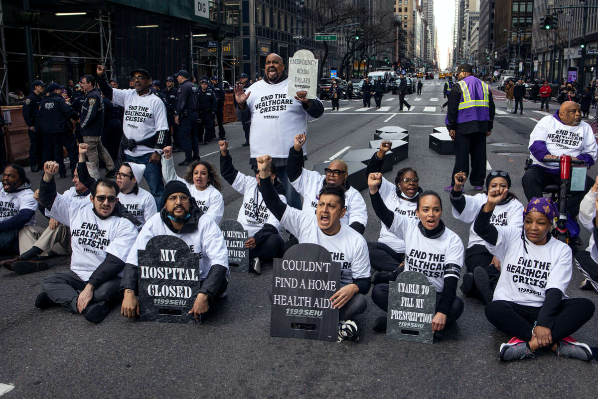 Protesters holding headstone-shaped signs bearing statements like "COULDN'T FIND A HOME HEALTH AID" and "MY HOSPITAL CLOSED" as they block an intersection during an outdoor demonstration