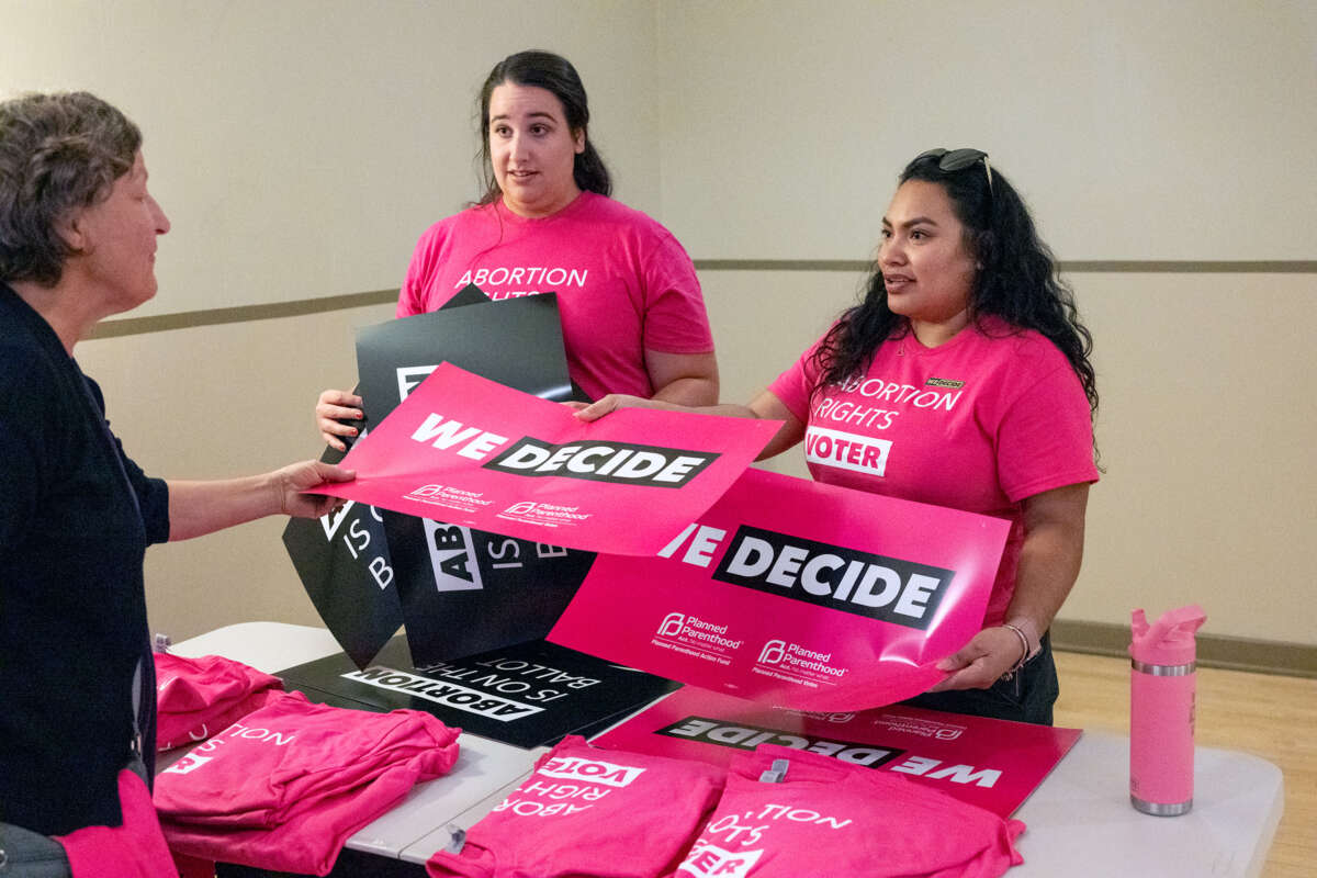 Two election workers in pink shirts that read "ABORTION RIGHTS VOTER" hand a pink sign reading "WE DECIDE" to someone they're speaking to across a table