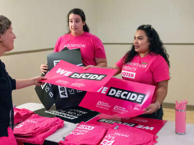 Two election workers in pink shirts that read "ABORTION RIGHTS VOTER" hand a pink sign reading "WE DECIDE" to someone they're speaking to across a table