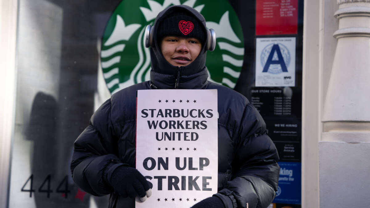 A smiling worker holds a sign reading "STARBUCKS WORKERS UNITED ON ULP STRIKE" during an outdoor action