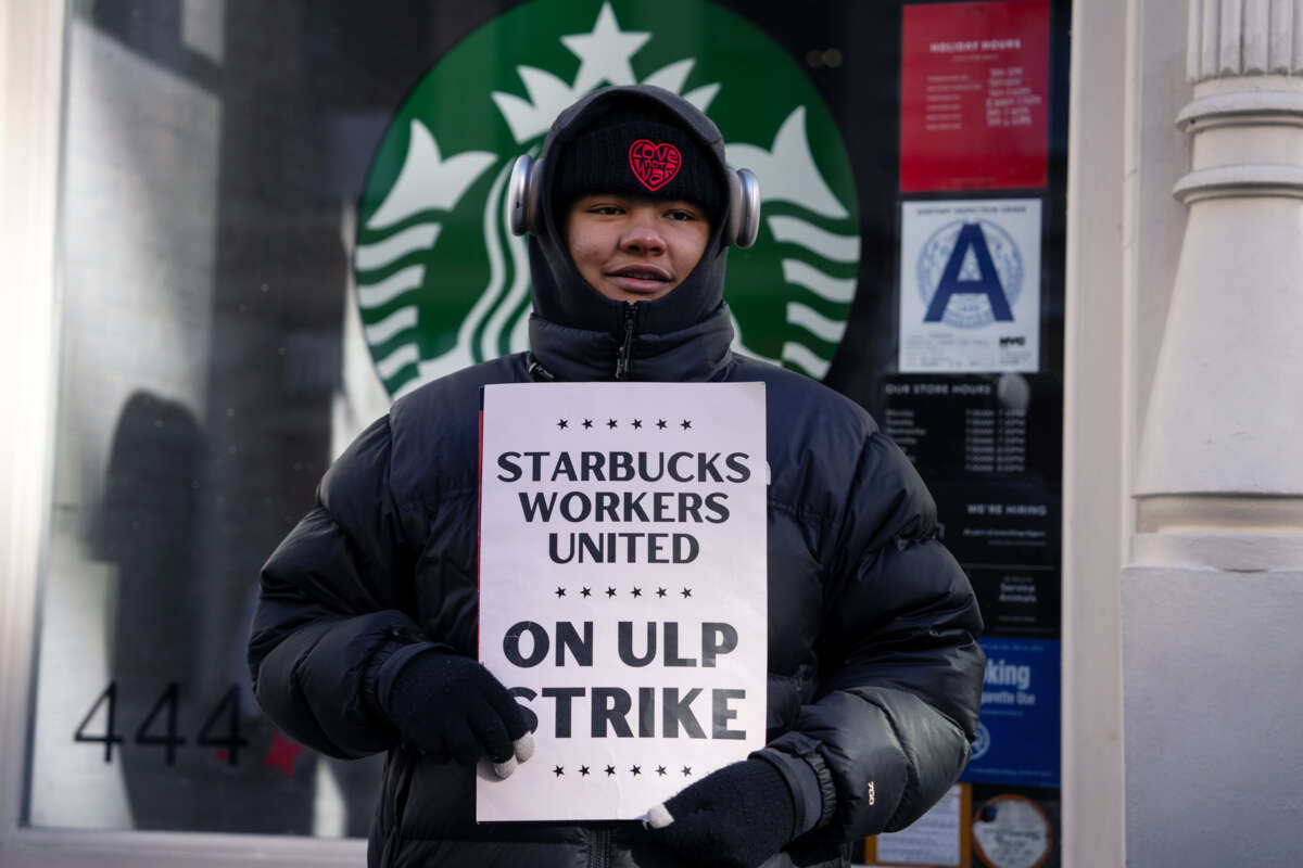 A smiling worker holds a sign reading "STARBUCKS WORKERS UNITED ON ULP STRIKE" during an outdoor action