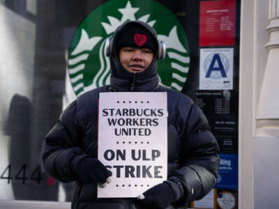 A smiling worker holds a sign reading "STARBUCKS WORKERS UNITED ON ULP STRIKE" during an outdoor action