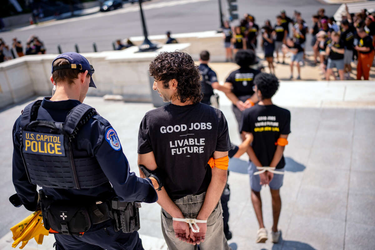 A protester wearing a shirt reading "GOOD JOBS; LIVEABLE FUTURE" and with hands zip-tied behind their back, is led away by a cop