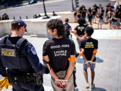 A protester wearing a shirt reading "GOOD JOBS; LIVEABLE FUTURE" and with hands zip-tied behind their back, is led away by a cop