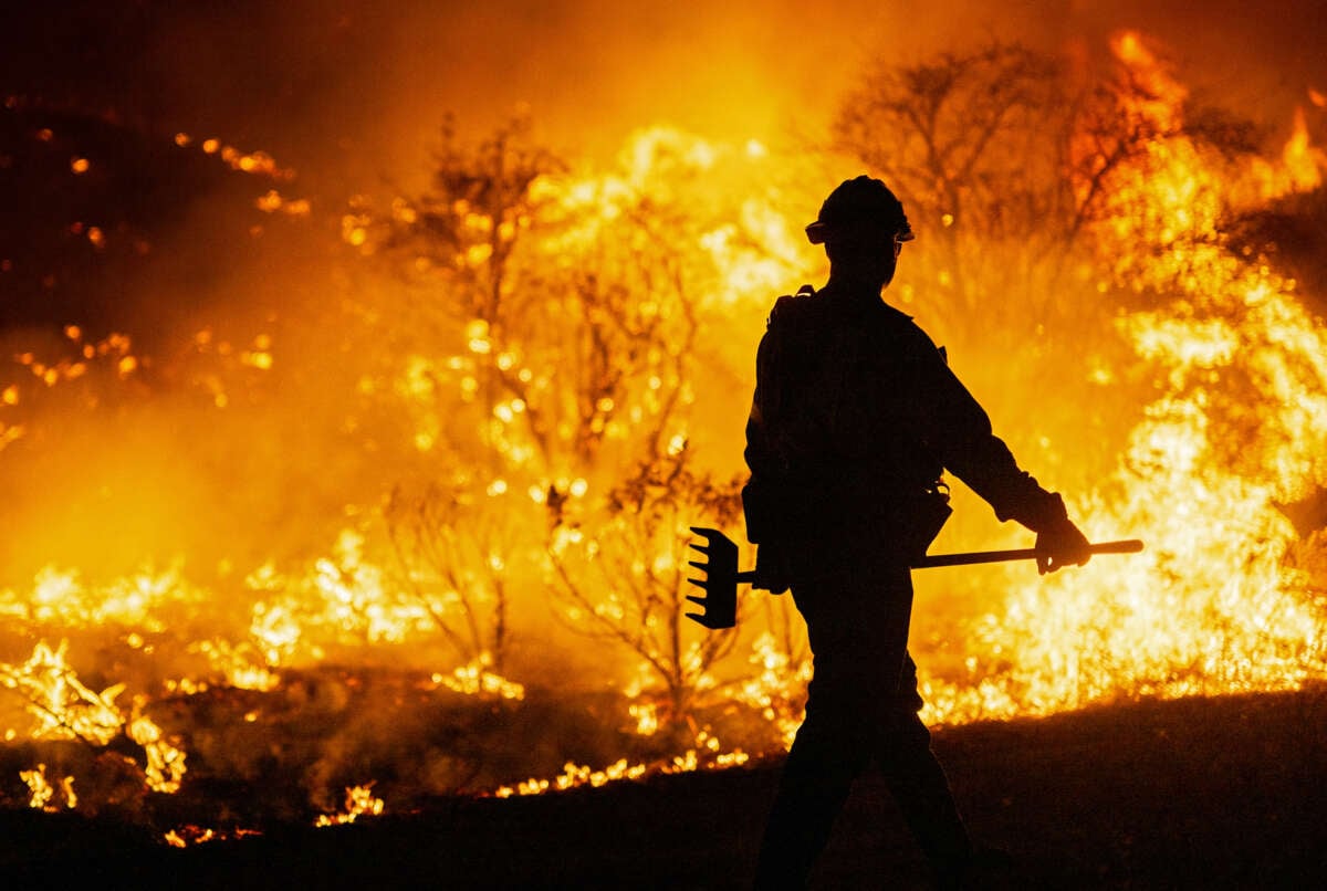 A firefighter holding a rake walks toward the flames surrounding him